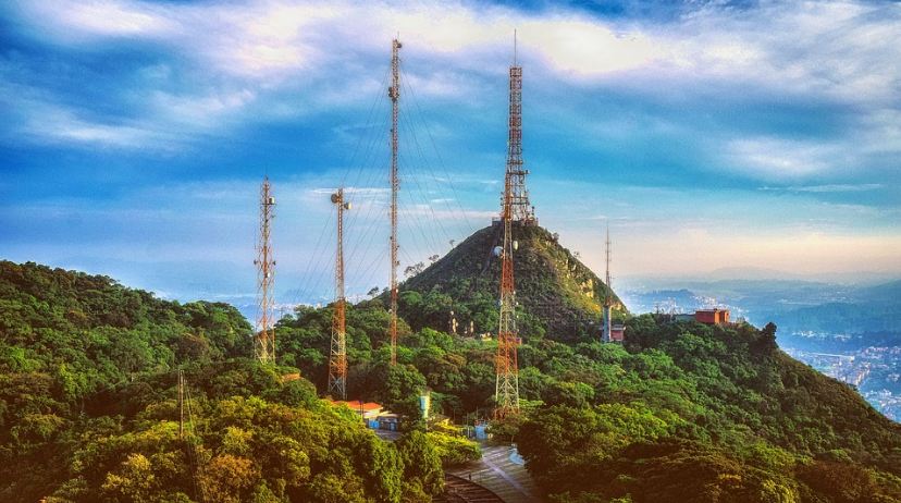 several cell towers, a tower on the top of a hill, trees, hills, area covered by trees, standing towers